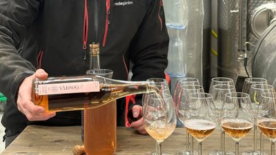 Mann mit Mütze hinter einem Tresen schenkt schäumendes Getränk aus einer Flasche in Gläser.