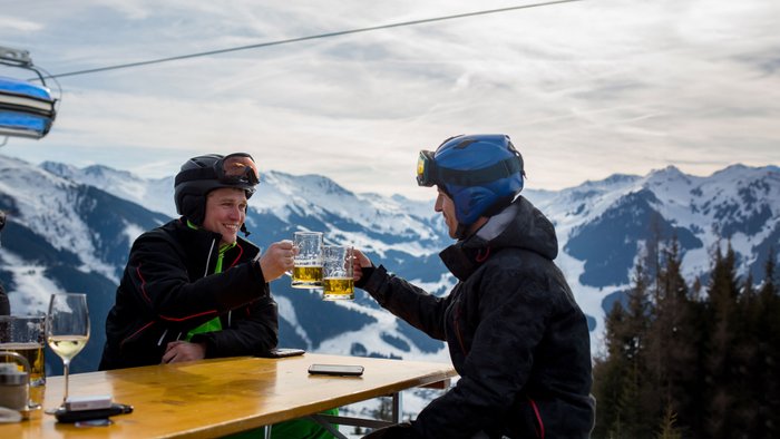 Zwei Skifahrer sitzen auf der Terrasse einer Skihütte und stoßen mit Bier an.