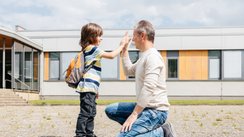 Ein Kind und sein Vater klatschen ab, im Hintergrund ein Schulgebäude