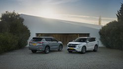 Ein grauer und ein weißer Mitsubishi Outlander stehen in der Abendsonne vor einem flachen Gebäude.