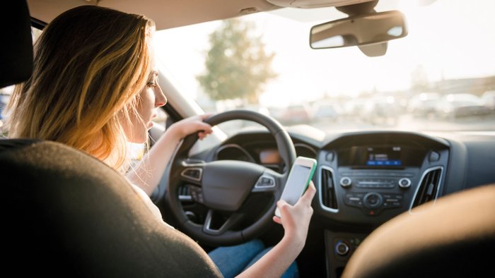 Eine Frau sitzt am Steuer eines Pkw und schaut auf ihr Smartphone.