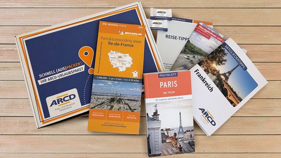 Ein ARCD-Urlaubspaket, daneben Reiseführer und Broschüren über Paris auf Holz.