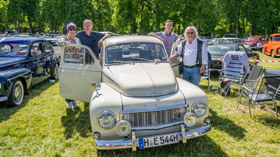 Oldtimer mit geöffneten Türen, daneben vier Personen. Im Hintergrund sind viele weitere Oldtimer zu sehen.