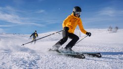 Zwei Skifahrer fahren bei blauem Himmel einen Skihang hinunter.