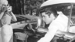 Elvis Presley lehnt sich aus einem Fahrzeugfenster und wird dabei von zwei Frauen fotografiert.