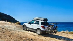 Ein Pkw mit Dachbox und Fahrrädern auf einem Fahrradträger vor einer Küstenkulisse.