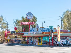Blick auf ein Burger-Restaurant am Rand einer Straße mit bunten Schildern, Möbeln und Classic Cars.