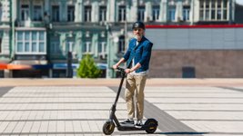 Mann mittleren Alters mit Sonnebrille und Helm fährt einen E-Scooter in einer Stadt.