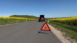 Ein Auto steht mit einer Panne am Straßenrand, im Vordergrund ein Warndreieck.
