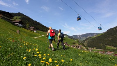Paar wandert in den Bergen über Blumenwiese, im Hintergrund Gondelbahn.