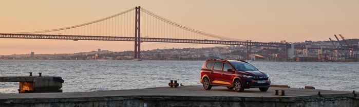 Ein roter Dacia Jogger Hybrid steht am Ufer bei Sonnenuntergangs-Stimmung. 