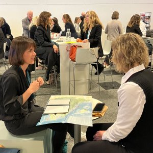 Zwei Frauen sitzen im Gespräch an einem Tisch und haben eine Landkarte vor sich, im Hintergrund andere Personen im Gespräch.