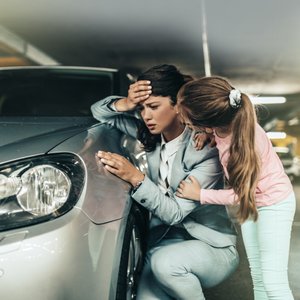 Kind guckt ob es Frau gut geht, welche sich den Kopf hält und am Auto abstützt.