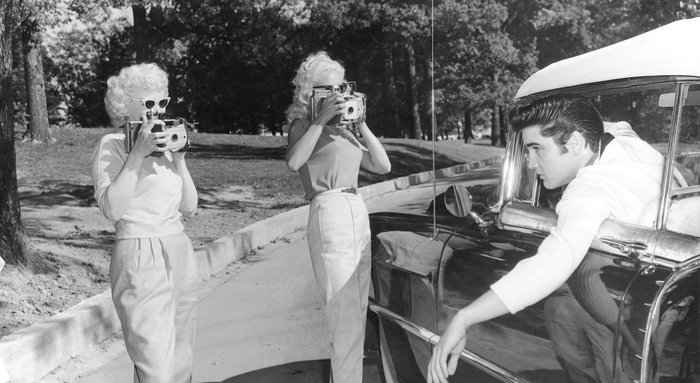 Elvis Presley lehnt sich aus einem Fahrzeugfenster und wird dabei von zwei Frauen fotografiert.