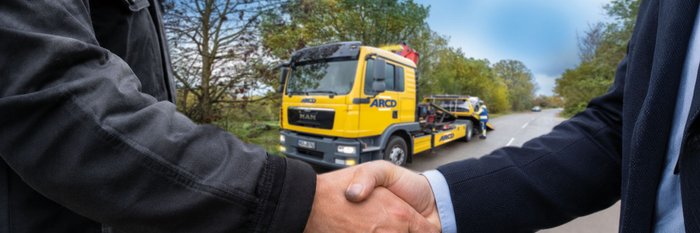 2 Personen schütteln sich die Hände, im Hintergrund wird ein Pkw abgeschleppt.