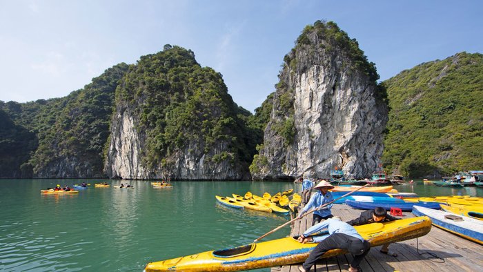 Männer ziehen auf einem Steg ein Boot an Land, im Hintergrund viele weitere Boote im türkisblauen Wasser und hohe begrünte Felsen.