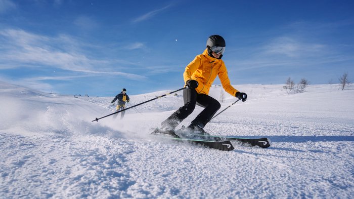 Zwei Skifahrer fahren bei blauem Himmel einen Skihang hinunter.