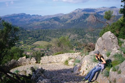 Eine junge Frau sitzt auf einer Steinmauer und blickt auf eine mediterrane Berglandschaft.