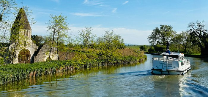 Hausboot fährt in idyllischer, grüner Landschaft an Kirchenruine vorbei.