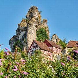 Blumen, Fachwerk-Burg und hochaufragender Nadel-Fels im Hintergrund.