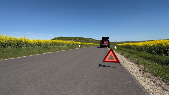 Ein Auto steht mit einer Panne am Straßenrand, im Vordergrund ein Warndreieck.