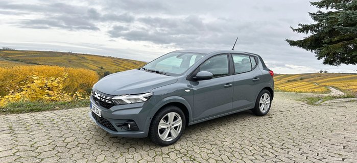 Ein graues Auto auf einem Parkplatz, im Hintergrund Felder und Wiese.