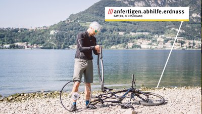 Ein Fahrradfahrer hat einen platten Reifen. Die 3-Wort-Adresse "anfertigen.abhilfe.erdnuss" steht in weißen Feld.
