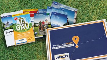 Ein ARCD-Urlaubspaket, daneben Reiseführer und Broschüren auf grünem Rasen.