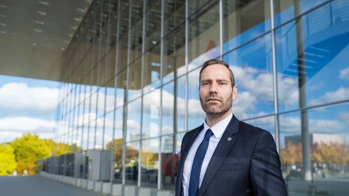 Ein Mann mit ernster Miene steht vor einem Gebäude mit Glasfassade.