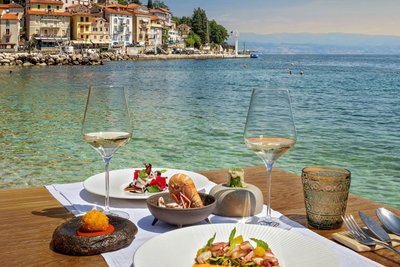 Gedeckter Tisch mit Tellern und Weingläsern am Meer mit Küstenort im Hintergrund.