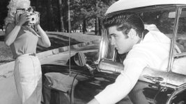 Elvis Presley lehnt sich aus einem Fahrzeugfenster und wird dabei von zwei Frauen fotografiert.