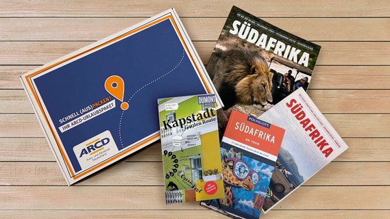Ein ARCD-Urlaubspaket, daneben Reiseführer und Broschüren über Südafrika auf Holz.