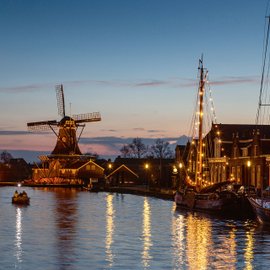 Abendlicher Blick auf einen Fluss mit beleuchteten Booten und Windmühle.