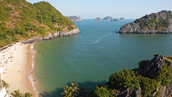 Blick aus der Vogelperspektive auf einen Sandstrand mit türkisblauem Wasser, der von begrünten Hügeln umrahmt wird.