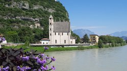 Blick auf den Fluss Etsch in Trento mit Blumen im Vordergrund und einer Kirche im Hintergrund.