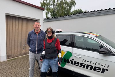 Fahrschülerin im Altersanzug mit dem Fahrlehrer vor dem Fahrschulauto.