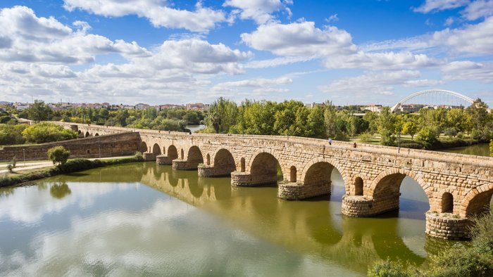 Landschaft mit römischer Steinbrücke über Fluss und moderner Stahlbrücke im Hintergrund.
