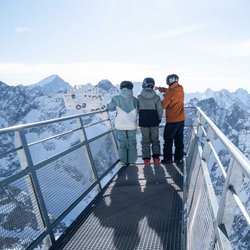Drei Skifahrer stehen auf einem Aussichtssteg aus Stahl und genießen den Blick auf die verschneiten französischen Südalpen.