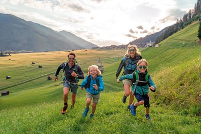 Vater, Mutter und zwei Kinder rennen fröhlich über eine grüne Wiese mit Bergen im Hintergrund.