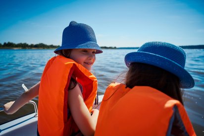 Zwei Kinder mit orangefarbenen Rettungswesten sitzen in einem Boot.