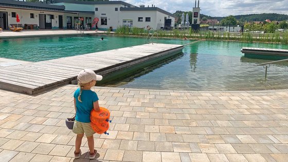 Ein Kind steht mit Schwimmflügeln vor einem Badesee.
