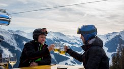 Zwei Skifahrer sitzen auf der Terrasse einer Skihütte und stoßen mit Bier an.