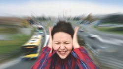 Frau hält sich die Ohren zu, im Hintergrund eine Straße mit Autos.