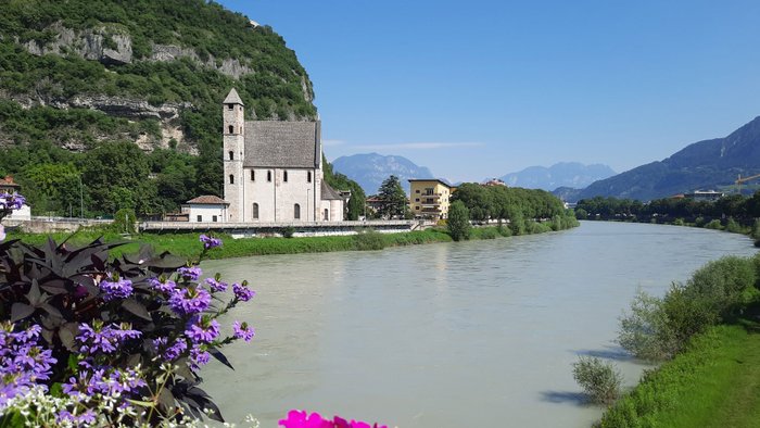 Blick auf den Fluss Etsch in Trento mit Blumen im Vordergrund und einer Kirche im Hintergrund.