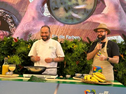 Zwei Männer stehen mit Kochgeschirr, Saft und Bananen an einem Pult vor buntem Hintergrund und bereiten Essen zu.