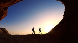 Zwei Wanderer – im Gegenlicht – laufen einen Felsbogen entlang.