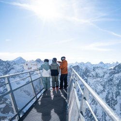 Drei Skifahrer stehen auf einem Aussichtssteg aus Stahl und genießen den Blick auf die verschneiten französischen Südalpen.