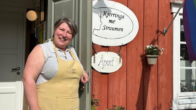 Lächelnde Frau mit Schürze schaut aus der Tür eines roten Holzhauses.