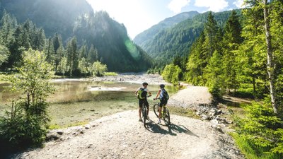 Zwei Mountainbiker halten an einem glasklaren See vor Bergkulisse.