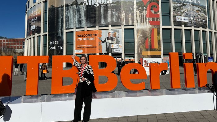 Frau steht vor ITB-Schriftzug, im Hintergrund Messeeingang mit bunten Werbeplakaten.